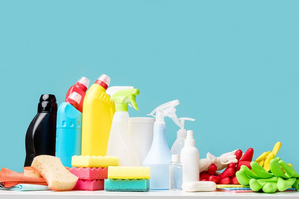 Assortment of colourful household cleaning bottles, sprays, sponges and gloves on a teal background