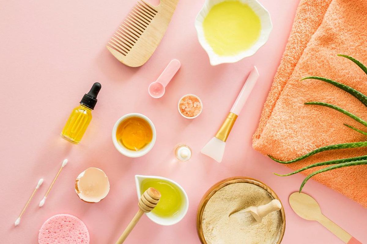 Flat lay of natural skincare and personal care ingredients including oils, aloe, clay and a comb on a pink background