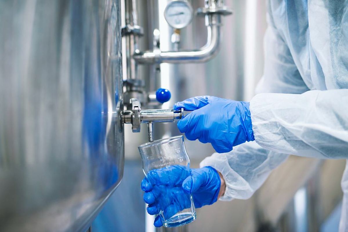 Laboratory technician in protective clothing and gloves drawing a water sample from a stainless-steel process tank at Mwembeshi Chemicals, Zambia
